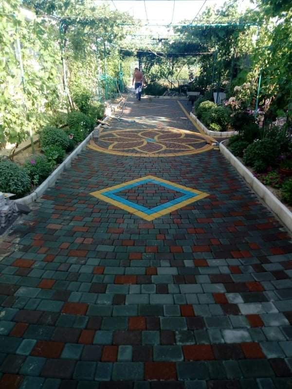 Kolorowa ścieżka z kostki brukowej w ogrodzie, z wzorem kwiatowym i rombem, prowadząca pod zielonym tunelem z roślin.