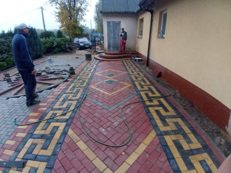 Układanie kostki brukowej w wzór grecki z czerwono-żółto-szarych kostek, widoczni dwaj pracownicy, jeden w trakcie pracy, drugi nadzoruje, w tle dom i samochód.