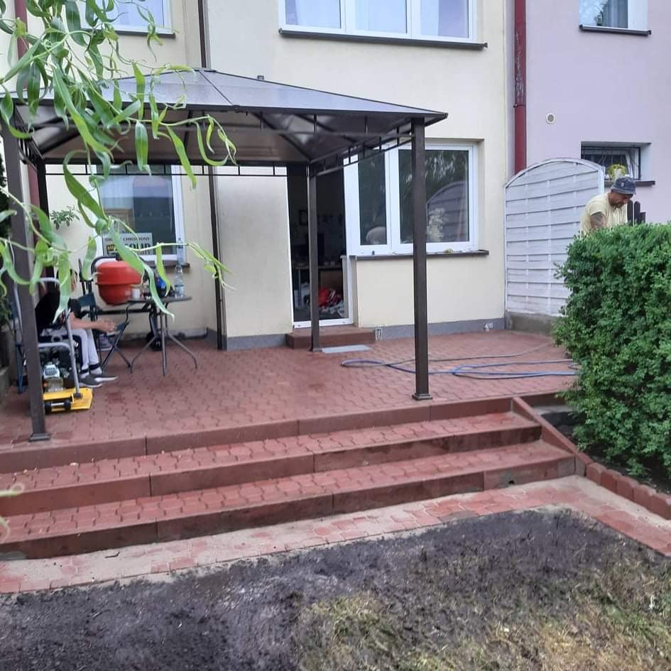 Czerwona kostka brukowa ułożona na tarasie z zadaszeniem przy jasnym domu, widoczny pracownik przycinający krzewy, betoniarka i zagęszczarka w tle, świeżo przygotowany teren pod trawnik.