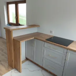 Szare szafki kuchenne z drewnianym blatem i płytą indukcyjną, widok na okno z drewnianą ramą i blat-barek. Podłoga wyłożona płytkami imitującymi marmur.