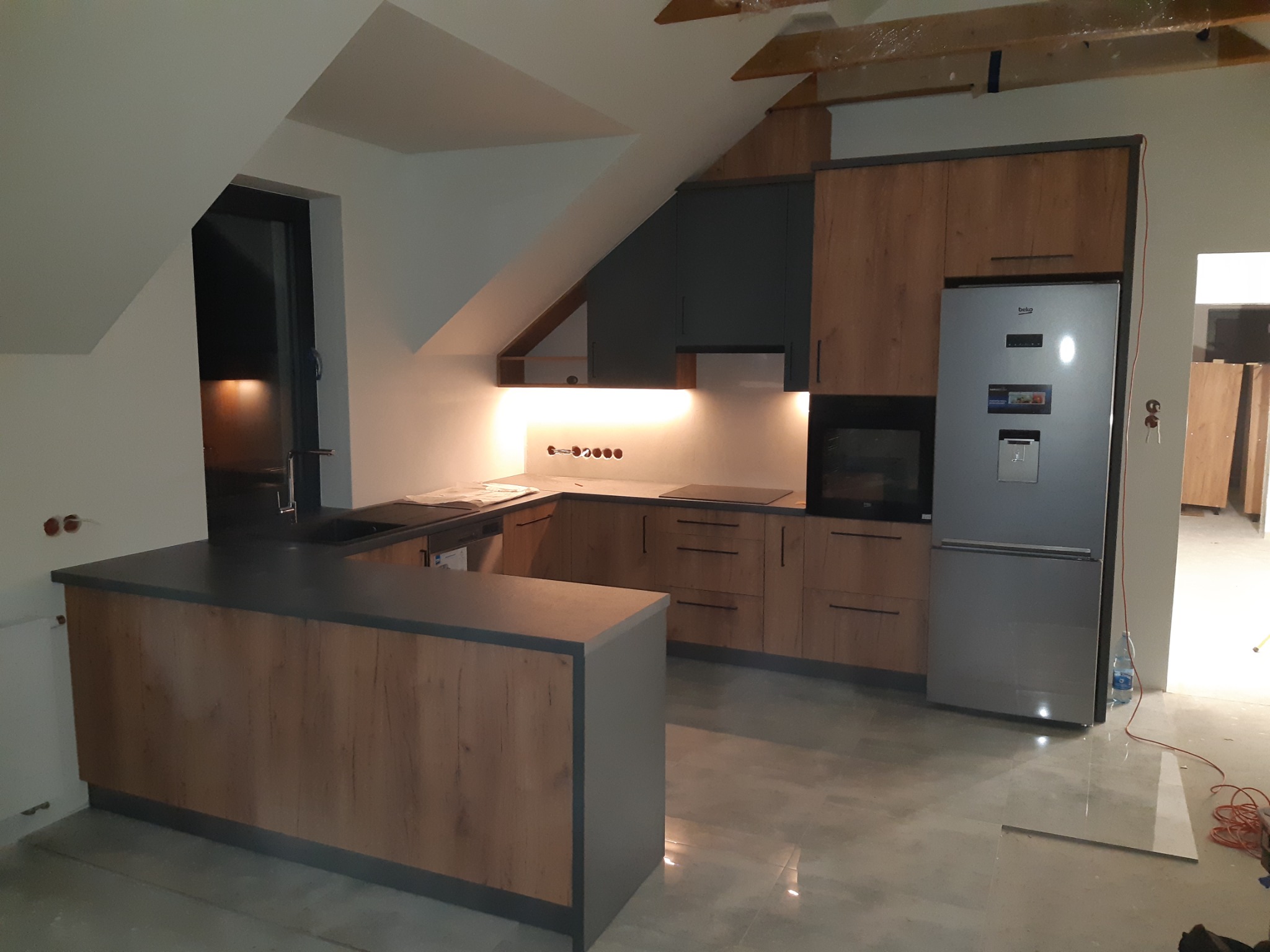 Nowoczesna kuchnia z drewnopodobnymi frontami i szarymi akcentami, zlokalizowana na poddaszu ze skośnym sufitem, oświetlona listwą LED pod szafkami górnymi, z widocznym fragmentem niezabezpieczonej...