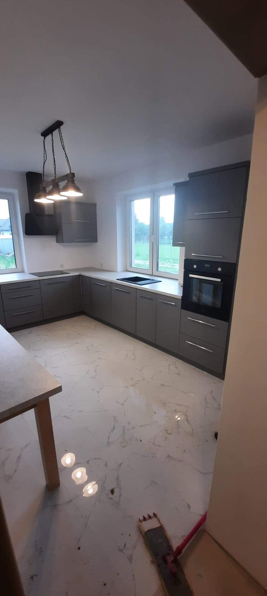 Szara kuchnia na wymiar z białym blatem i marmurową podłogą, oświetlona trzema lampami wiszącymi w stylu industrialnym, z widoczną czarną płytą indukcyjną i piekarnikiem.