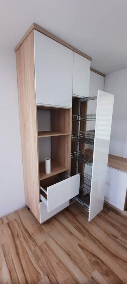 Wysoka szafka kuchenna z drewnopodobnymi bokami i białymi frontami, wyposażona w wysuwaną półkę cargo i otwartą półkę z rolką papieru. Widoczna drewniana podłoga.