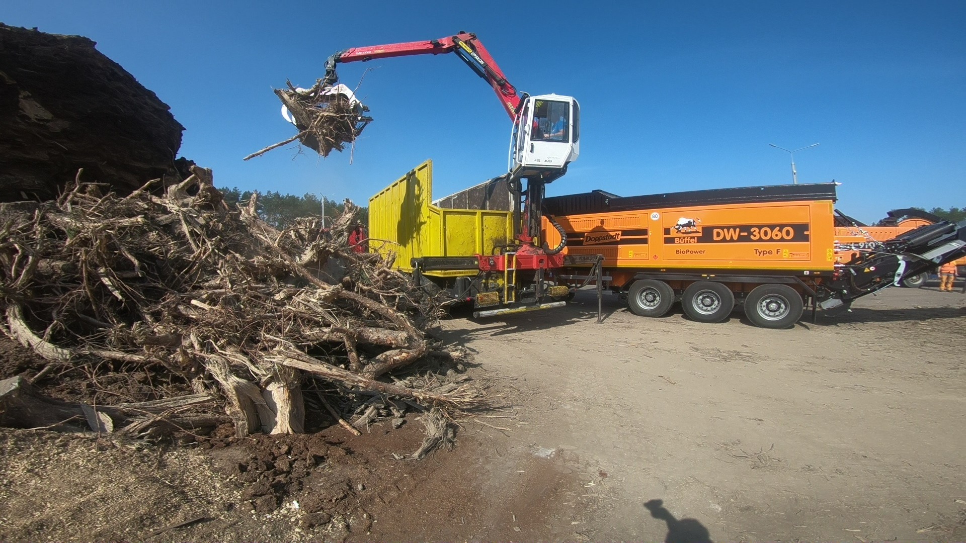 Ciężarówka z dźwigiem załadowuje korzenie drzew do żółtego kontenera. W tle sterta korzeni i maszyna do rozdrabniania.