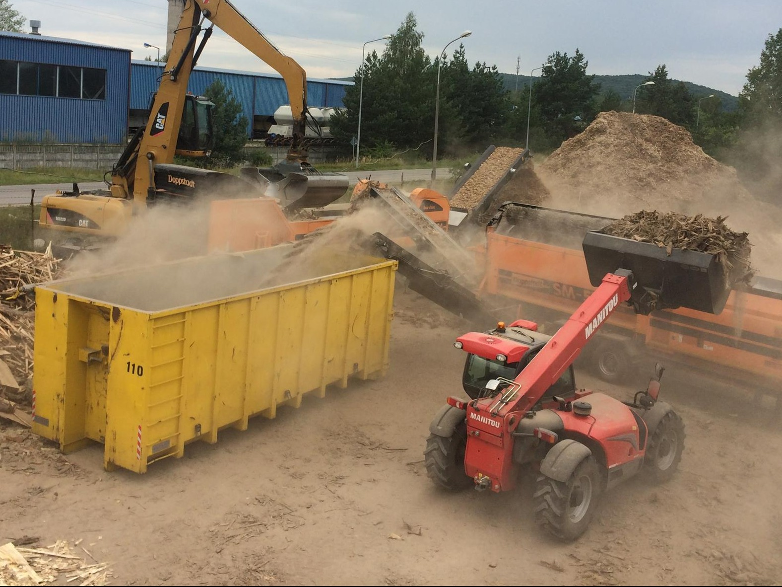 Przemysłowa obróbka drewna: załadunek zrębków do kontenera za pomocą ładowarki teleskopowej Manitou, w tle maszyna do rozdrabniania gałęzi i koparka.
