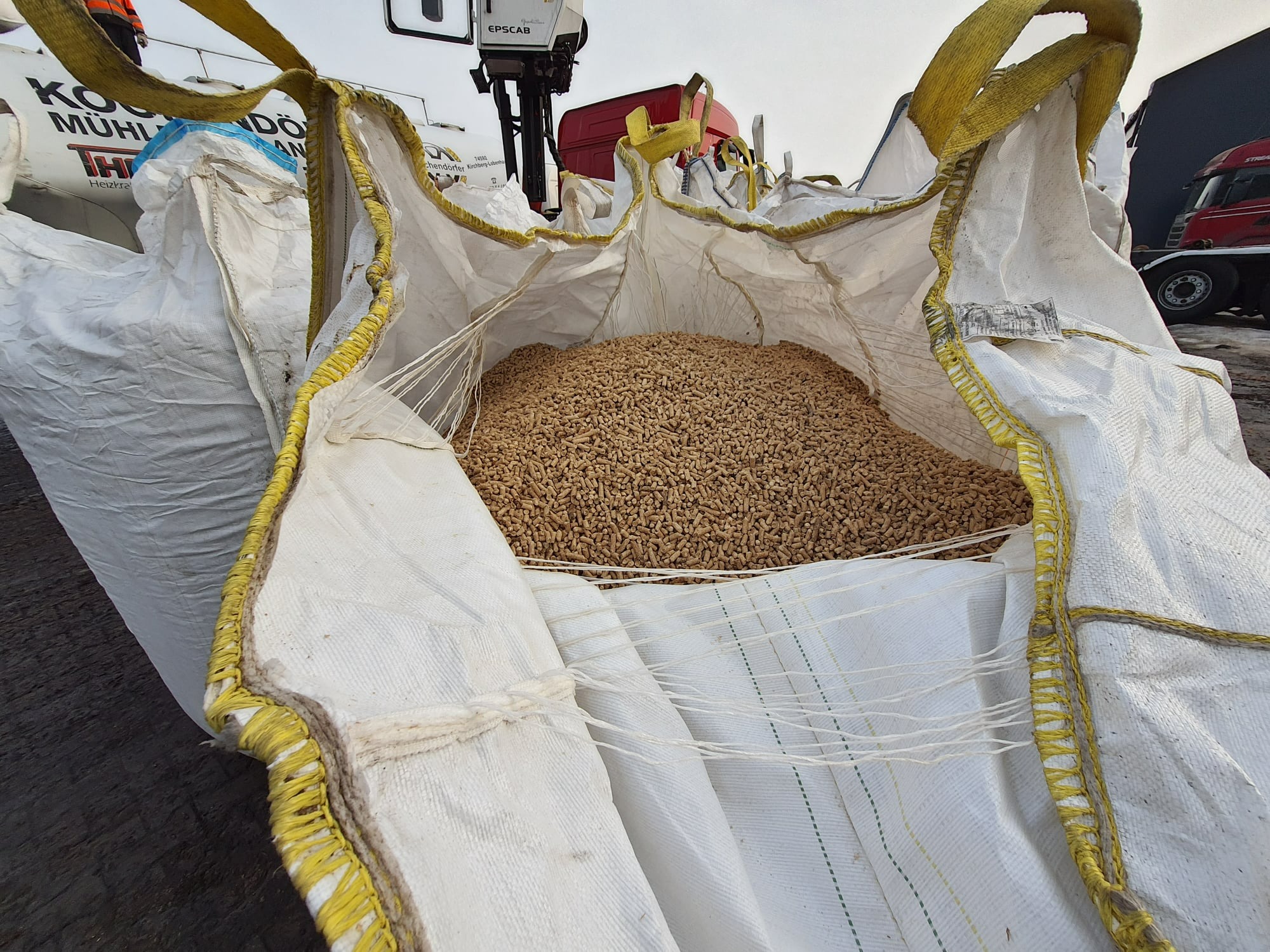 Wnętrze dużego, białego worka typu big bag wypełnionego jasnymi, brązowymi granulkami, prawdopodobnie paliwem stałym, z widocznymi żółtymi wzmocnieniami i fragmentami ciężarówki w tle.