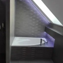 Nowoczesna łazienka na poddaszu z białą wanną, szarą glazurą w odcieniach szarości, oświetleniem LED podkreślającym skośny sufit i minimalistyczną armaturą.