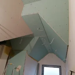 Skośny sufit poddasza w trakcie zabudowy płytami gipsowo-kartonowymi, widoczne kable oświetleniowe i drabina aluminiowa. Częściowo wyszpachlowane ściany i okno dachowe.