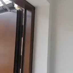 Fragment brązowej ościeżnicy drzwiowej z widocznym elementem montażowym, na tle świeżo pomalowanej, jasnoszarej ściany.