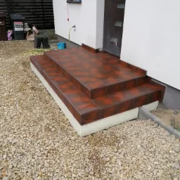 Wykończone, brązowe płytki na schodach wejściowych do domu, otoczone żwirem.