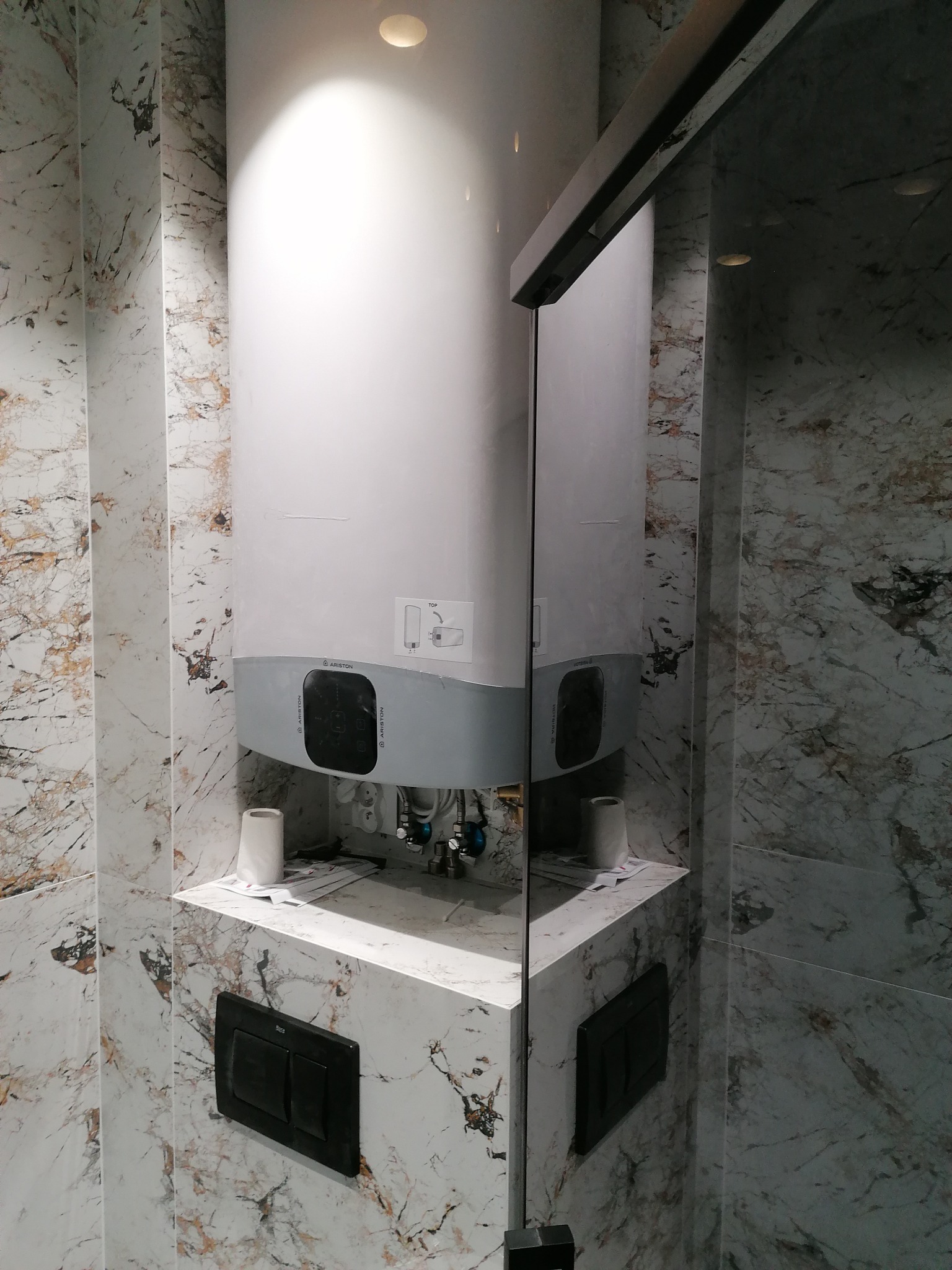 Biały podgrzewacz wody Ariston zamontowany w narożniku łazienki wykończonej płytkami imitującymi marmur, widoczny fragment kabiny prysznicowej i czarny panel spłuczki WC.
