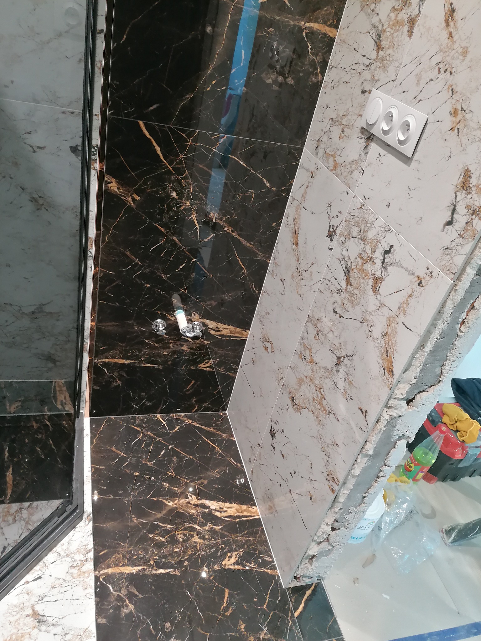 Łazienka w trakcie wykańczania: czarne i białe płytki imitujące marmur z widocznymi przyłączami wodnymi i puszką elektryczną.