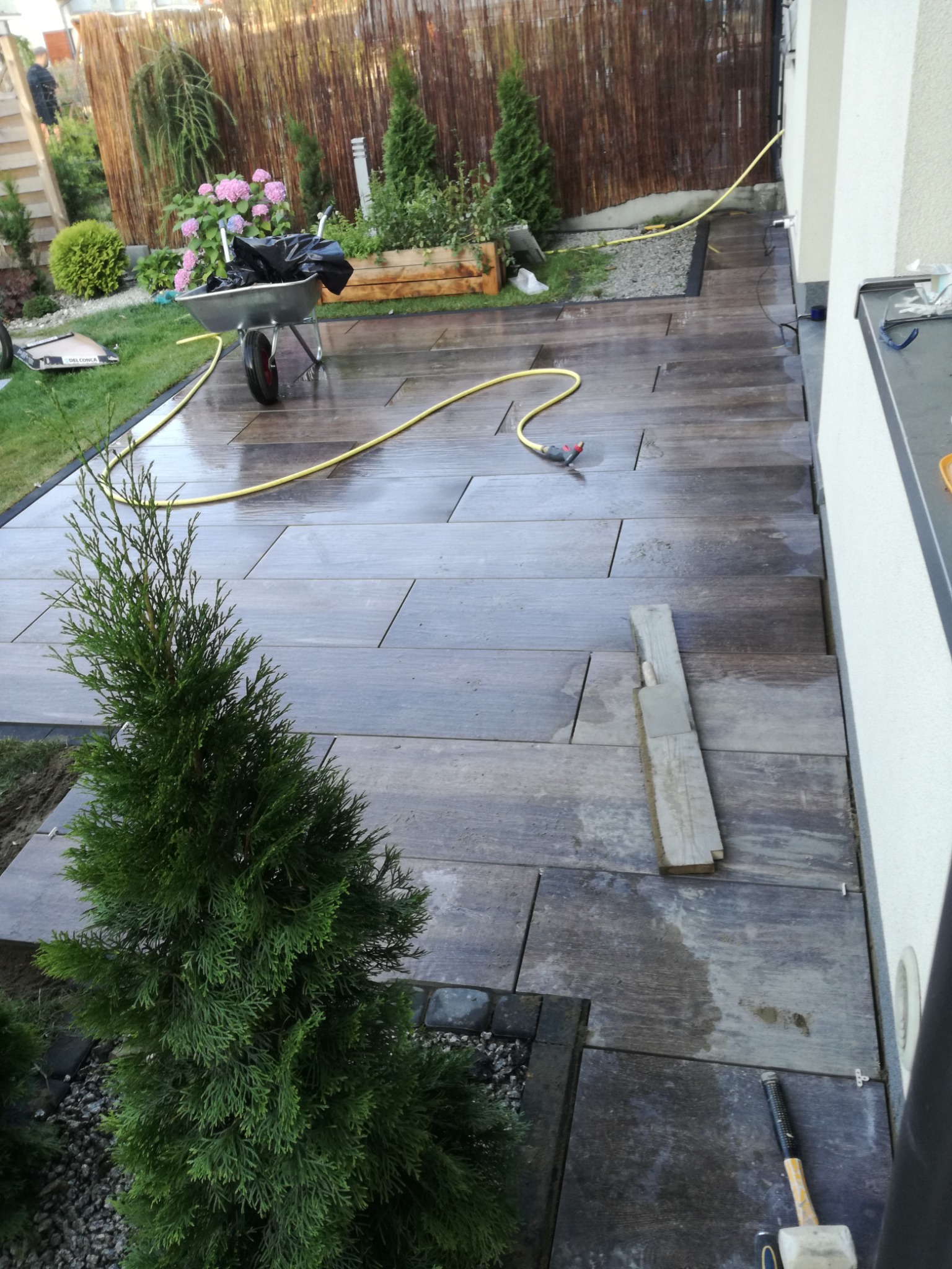 Układanie płytek imitujących drewno na zewnątrz budynku, widoczna poziomica, fuga, fragment trawnika i roślinności ogrodowej, w tle taczka z kwiatami.