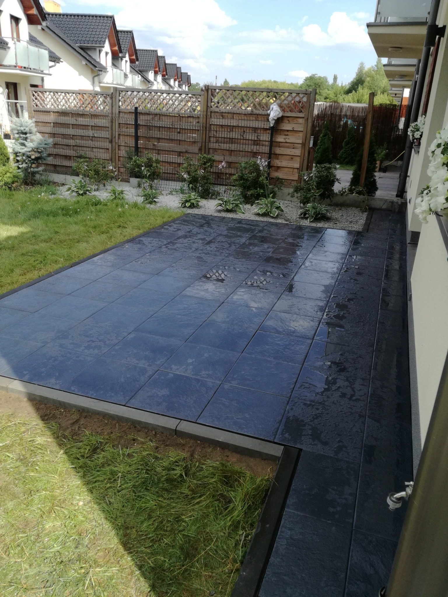 Nowoczesny taras wyłożony ciemnymi, kwadratowymi płytami, otoczony świeżą zielenią i drewnianym ogrodzeniem, z widocznymi śladami wody na powierzchni płytek.