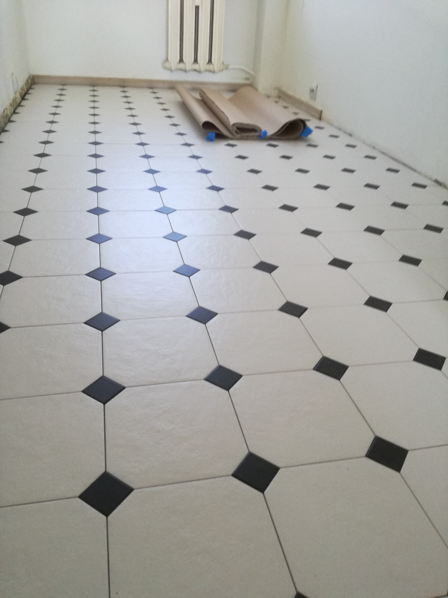 Wyłożona jasnymi płytkami podłoga z czarnymi, romboidalnymi akcentami; widoczne zabezpieczenie podłogi papierem pakowym w rogu pomieszczenia.