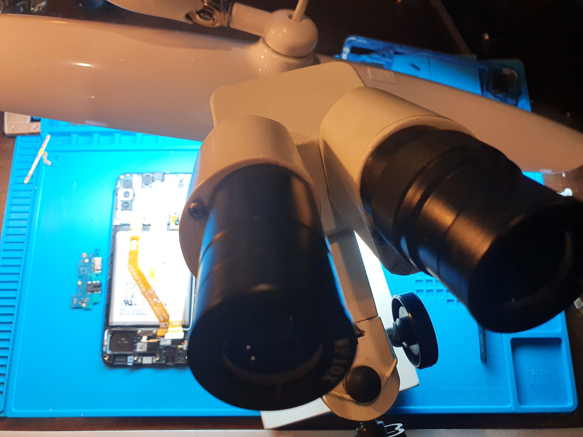 Mikroskop nad rozłożonym smartfonem na niebieskiej macie serwisowej, widoczne elementy elektroniki i bateria.