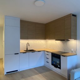 Masalon Design - Nowoczesna kuchnia w bieli i drewnie z czarną armaturą. Górne szafki z drewnianym fornirem, podświetlenie LED pod szafkami. Minimalistyczny design, jasne drewno na podłodze.