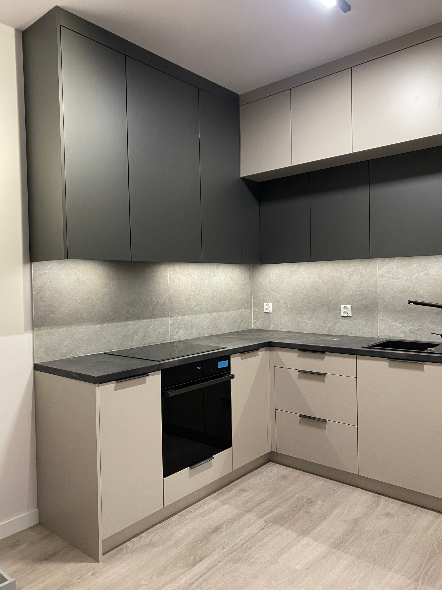 Nowoczesna kuchnia w odcieniach szarości i beżu z czarnymi akcentami. Górne szafki grafitowe, dolne beżowe. Blat z kamienia. Oświetlenie LED pod szafkami.