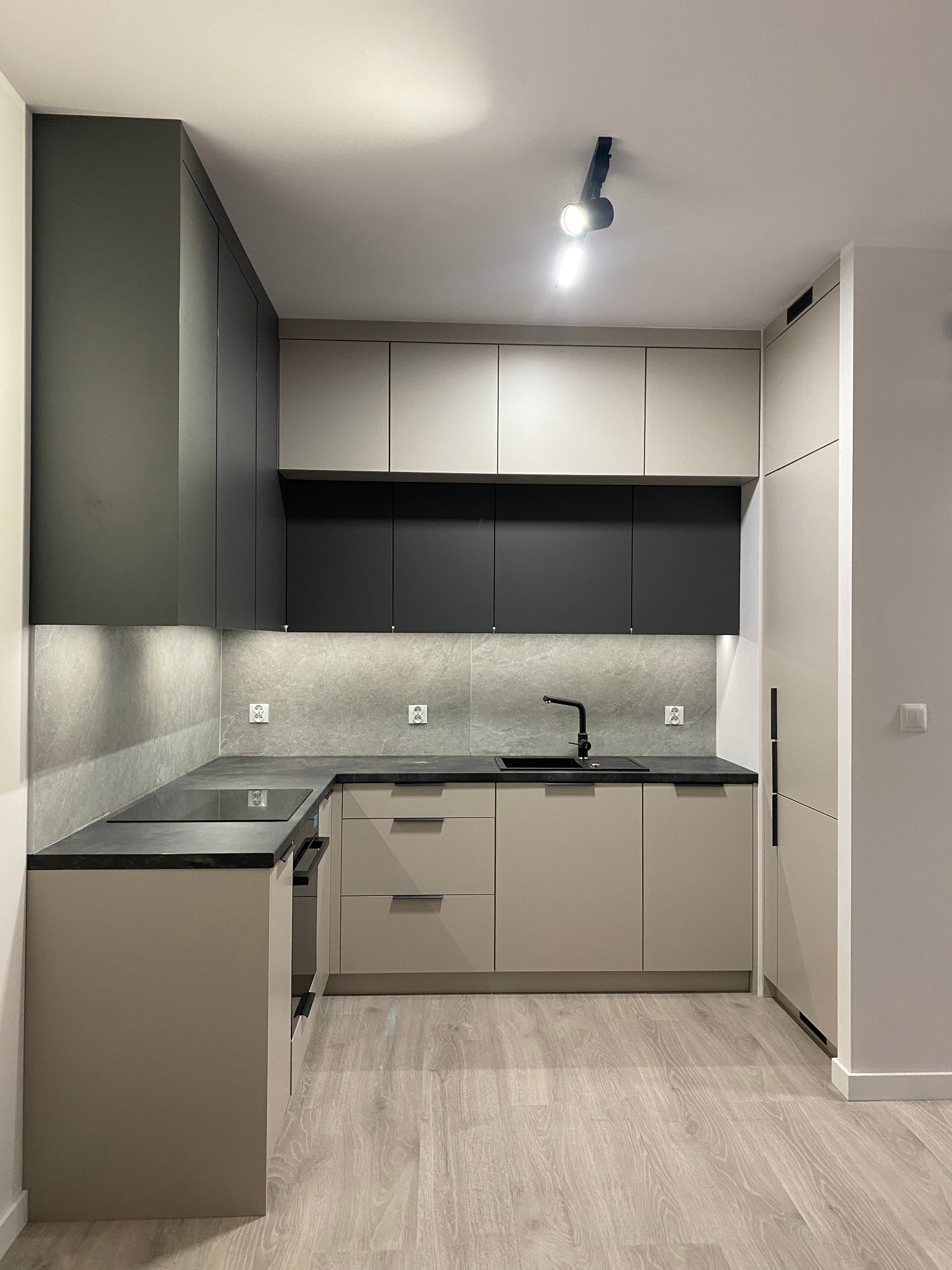 Nowoczesna kuchnia w odcieniach szarości i beżu z czarną armaturą. Minimalistyczny design, oświetlenie punktowe, drewnopodobna podłoga. Ujęcie z niskiego kąta.