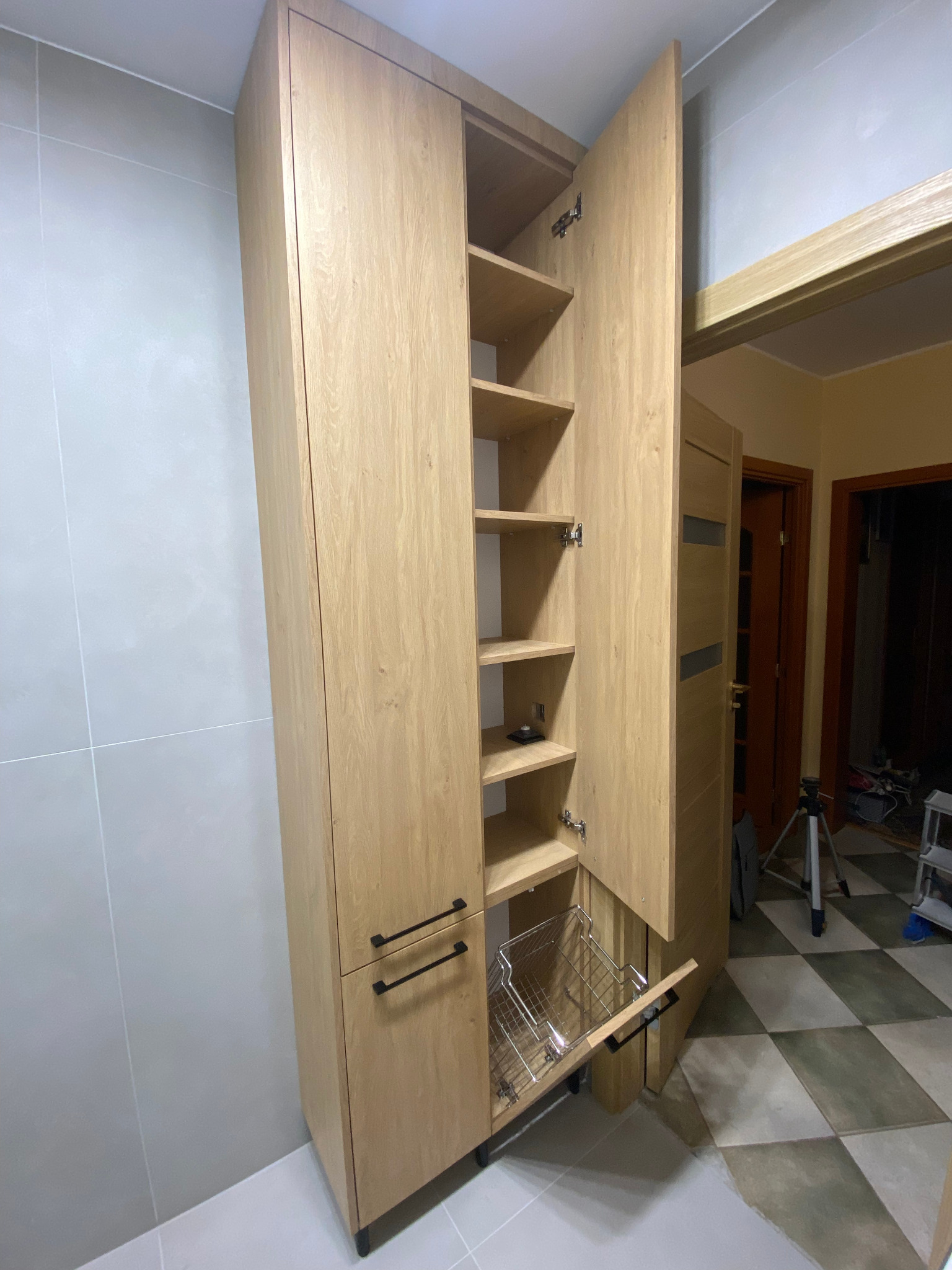 Wysoka, drewniana szafka łazienkowa z otwartymi drzwiami, ukazująca półki i wysuwany kosz na pranie. Jasne płytki na ścianie i podłodze. Widok na wnętrze mieszkania w tle.