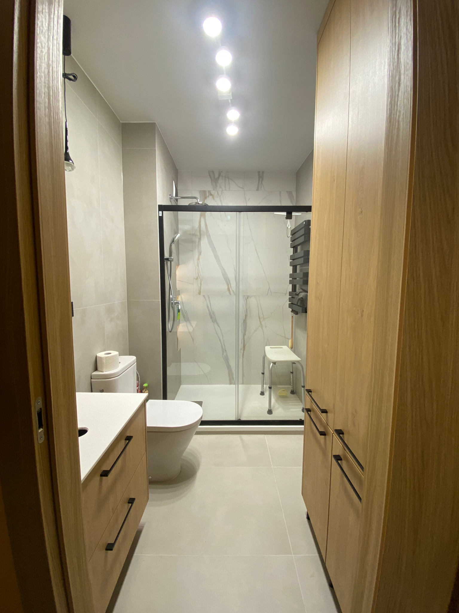 Nowoczesna łazienka z kabiną prysznicową typu walk-in, wykończona w jasnych odcieniach szarości i drewna. Uwagę zwraca minimalistyczny design i funkcjonalne szafki.