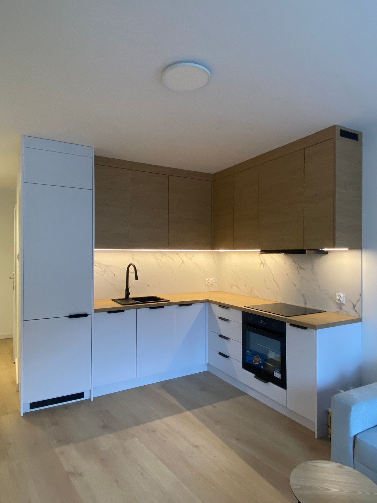 Nowoczesna kuchnia w bieli i drewnie z czarną armaturą. Górne szafki z drewnianym fornirem, podświetlenie LED pod szafkami. Minimalistyczny design, jasne drewno na podłodze.