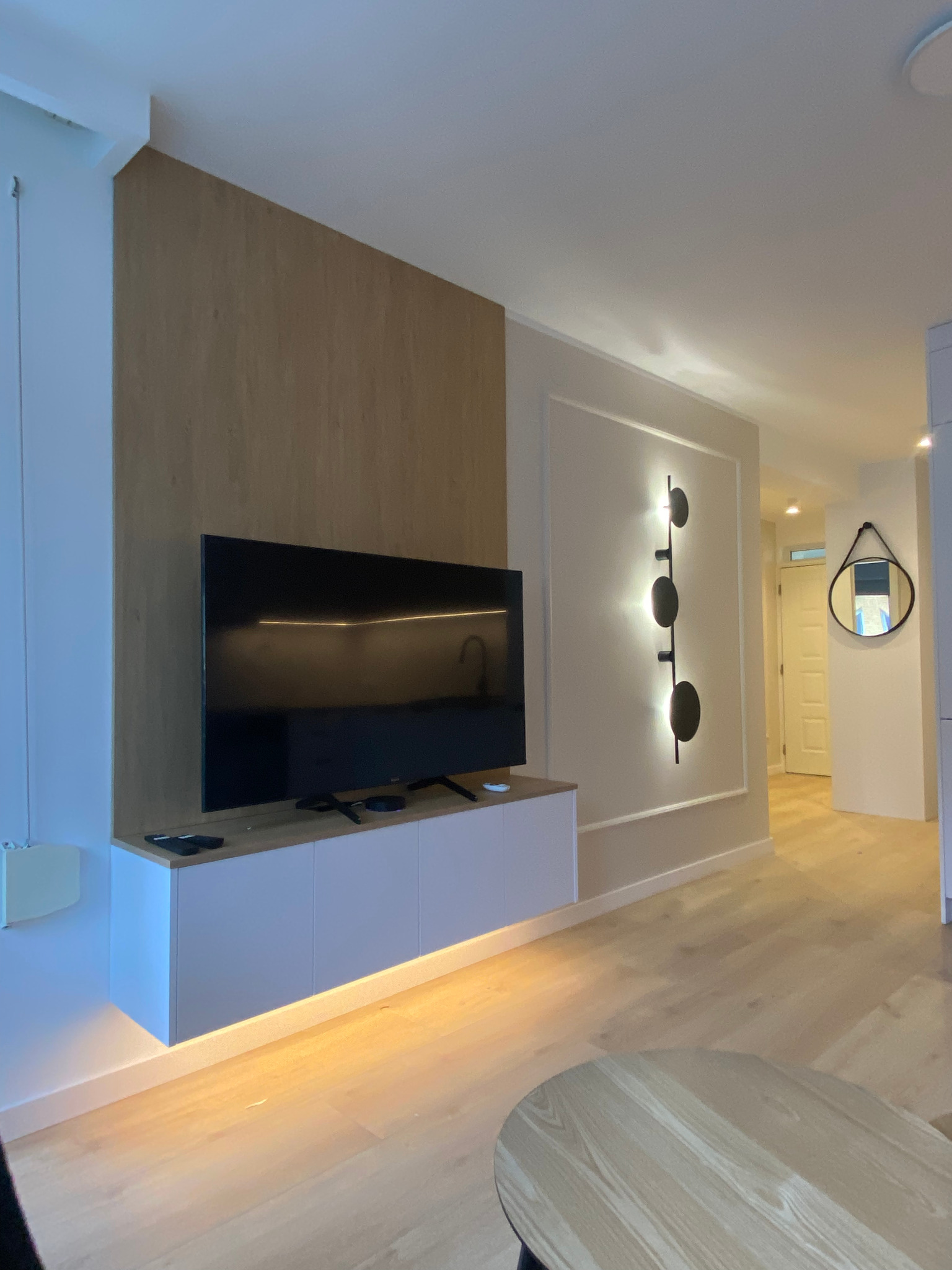 Nowoczesny salon z drewnianą ścianą, podświetlaną szafką RTV i designerską lampą ścienną. Lustro w okrągłej ramie w tle. Jasna podłoga i minimalistyczny wystrój.