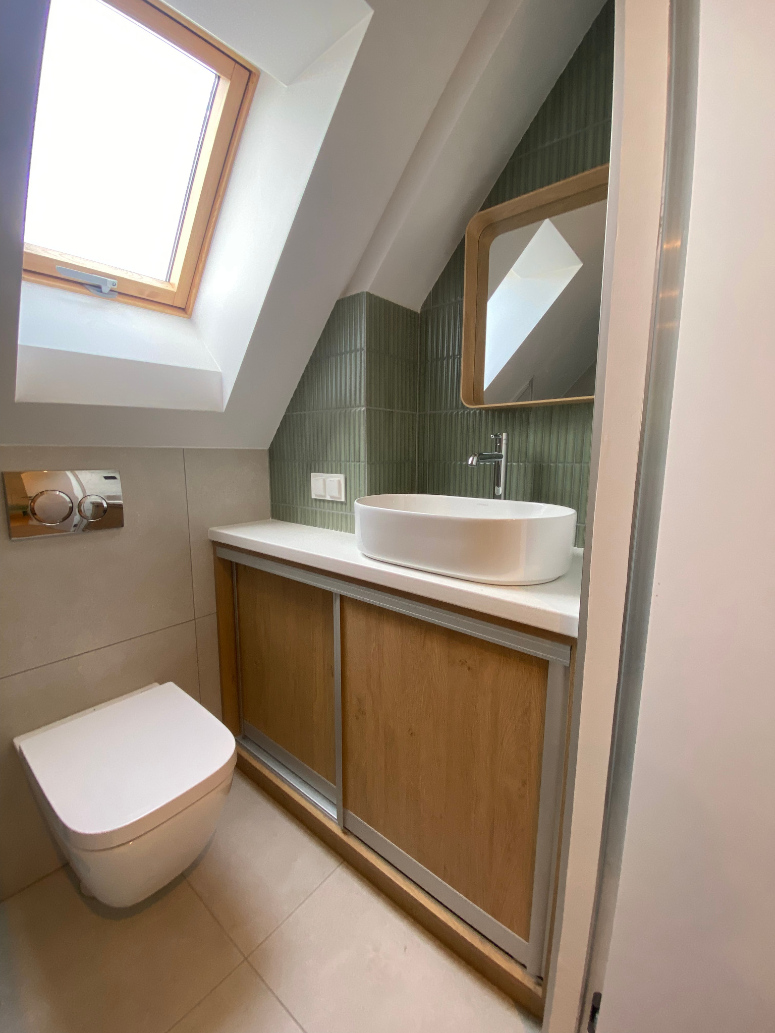 Nowoczesna łazienka na poddaszu z oknem dachowym, umywalką nablatową, lustrem w drewnianej ramie i toaletą podwieszaną. Użyto zielonej glazury i drewnianych frontów szafek.