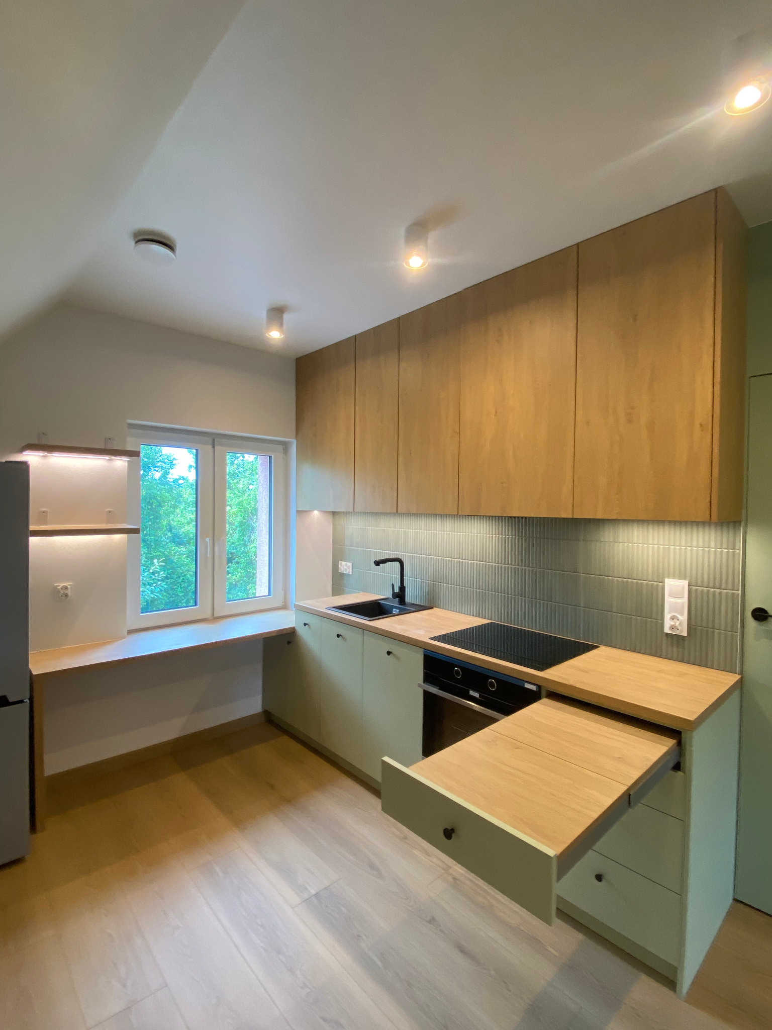 Jasna kuchnia z drewnianymi szafkami, wysuwanym blatem i oknem z widokiem na zieleń. Minimalistyczny design, oświetlenie punktowe i płytki w odcieniach szarości.