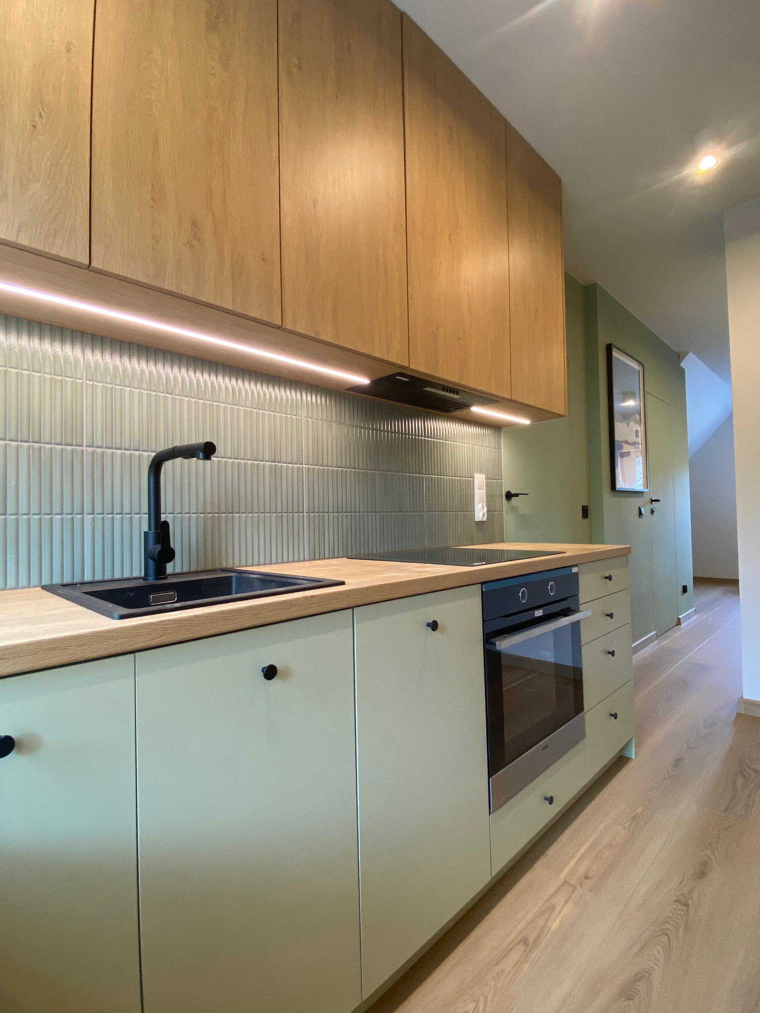 Nowoczesna kuchnia w odcieniach zieleni i drewna. Uwagę przykuwa czarna armatura i pionowe płytki nad blatem. Oświetlenie LED podkreśla minimalistyczny charakter.