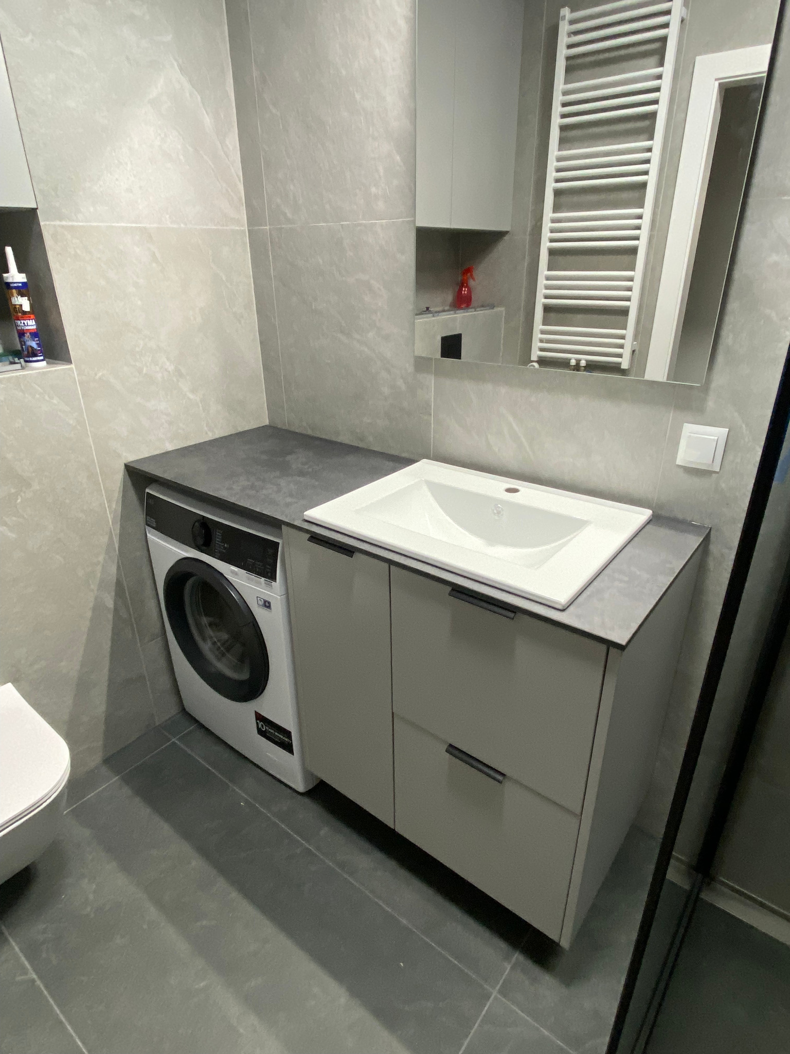 Nowoczesna łazienka w szarościach: pralka zabudowana pod blatem z umywalką, minimalistyczne szafki, grzejnik drabinkowy odbity w lustrze. Spokojna kolorystyka.