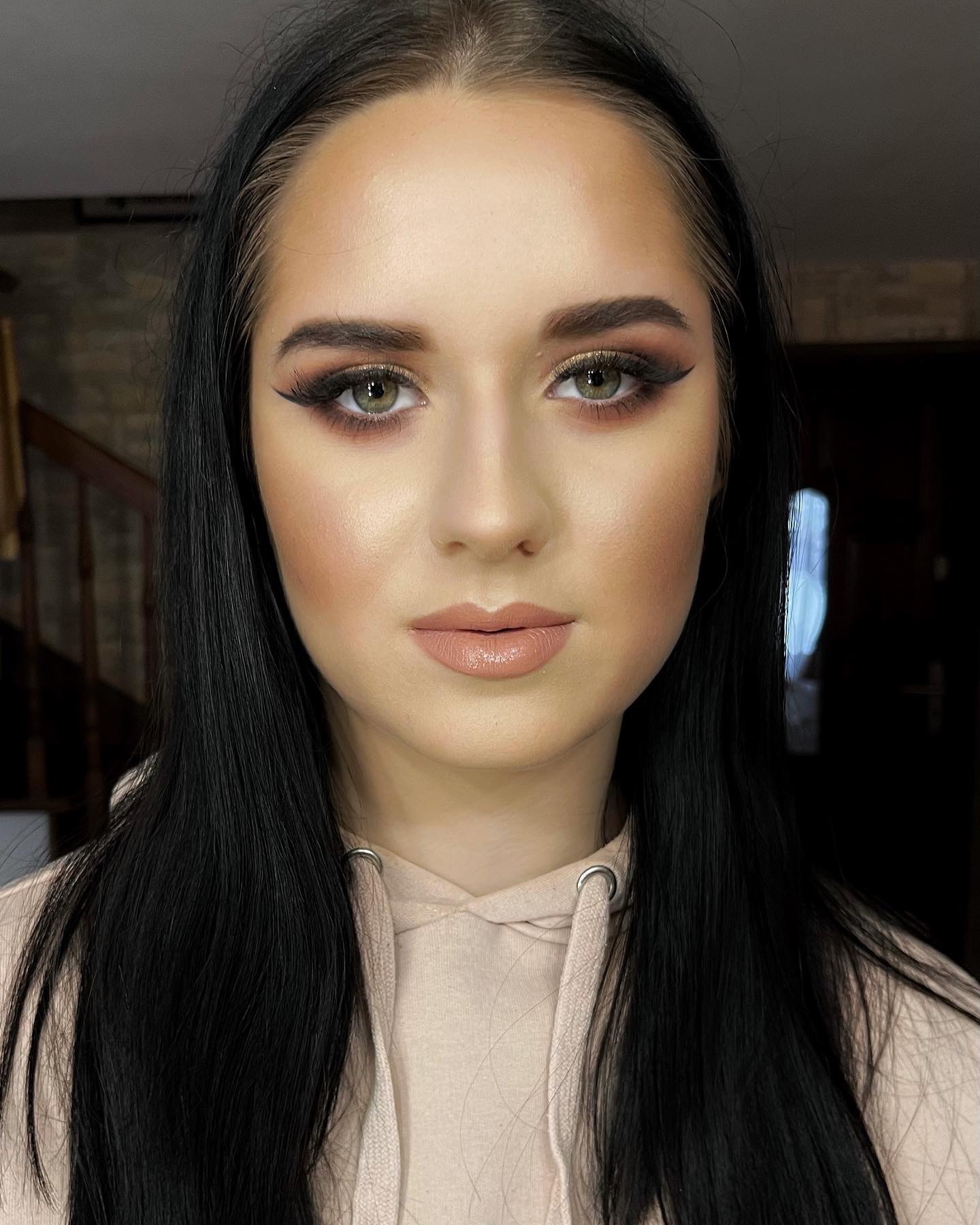 Portret kobiety z profesjonalnym wizażem: podkreślone brwi, złote cienie z czarną kreską, konturowanie policzków i błyszczące, beżowe usta, czarne, proste włosy opadające na beżową bluzę z kapturem.