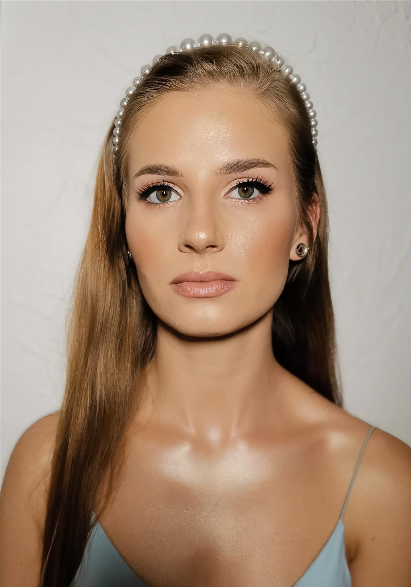 Portret kobiety z subtelnym, dziennym upiększeniem, podkreślonymi rzęsami i perłową opaską na włosach, na jasnym tle.