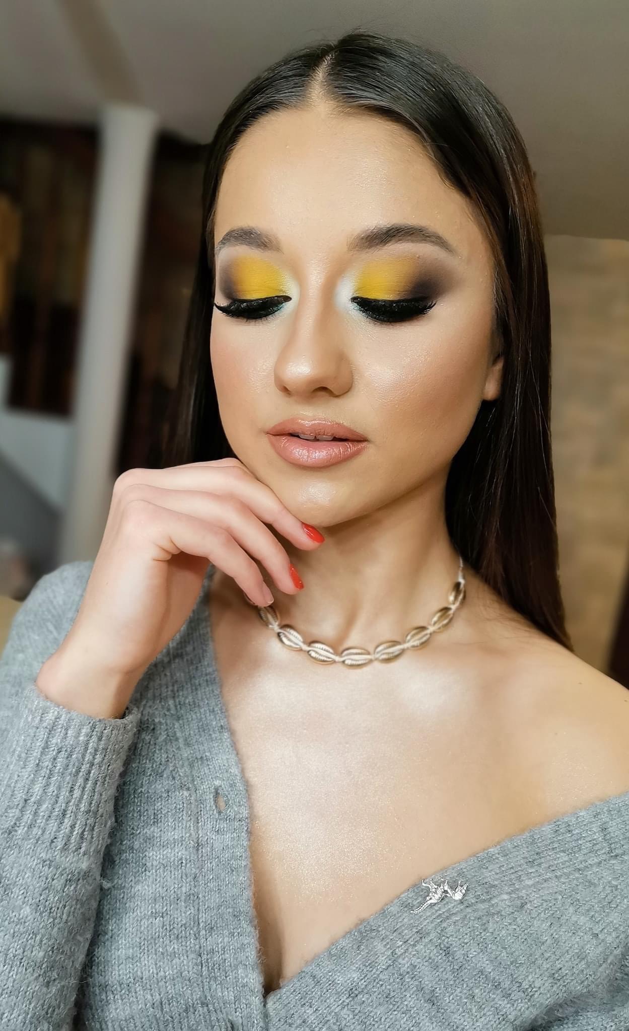 Portret młodej kobiety z intensywnym, żółto-czarnym cieniowaniem powiek i błyszczącymi ustami, dłoń podparta pod brodą, widoczny naszyjnik z muszelek i sweter z dekoltem.