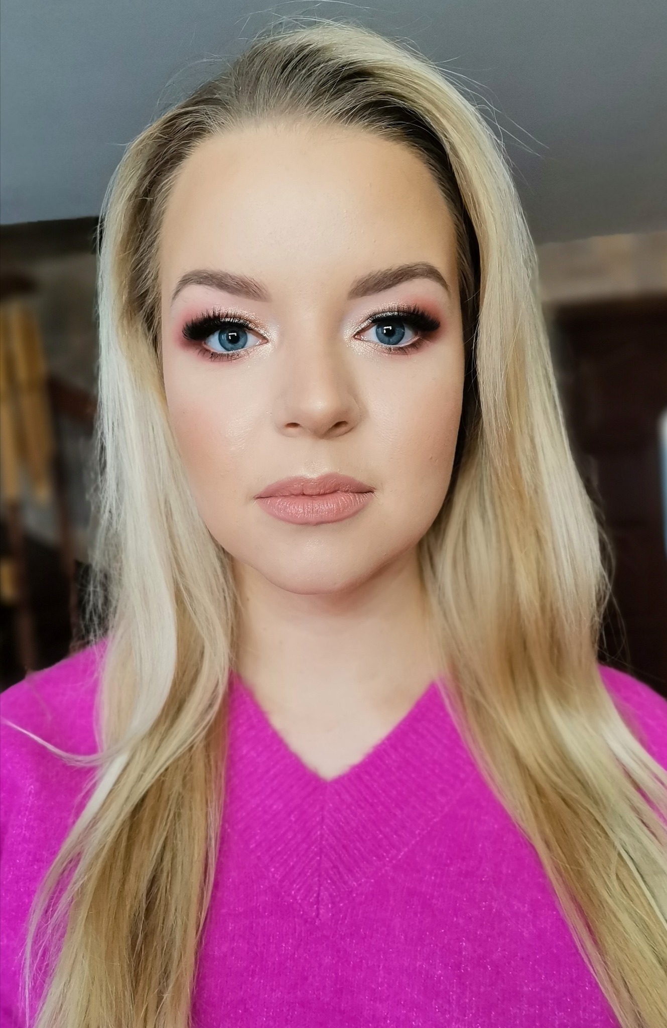 Portret blondynki z delikatnym, różowym cieniowaniem powiek i sztucznymi rzęsami, usta podkreślone błyszczykiem w odcieniu nude, ubrana w fuksjowy sweter.