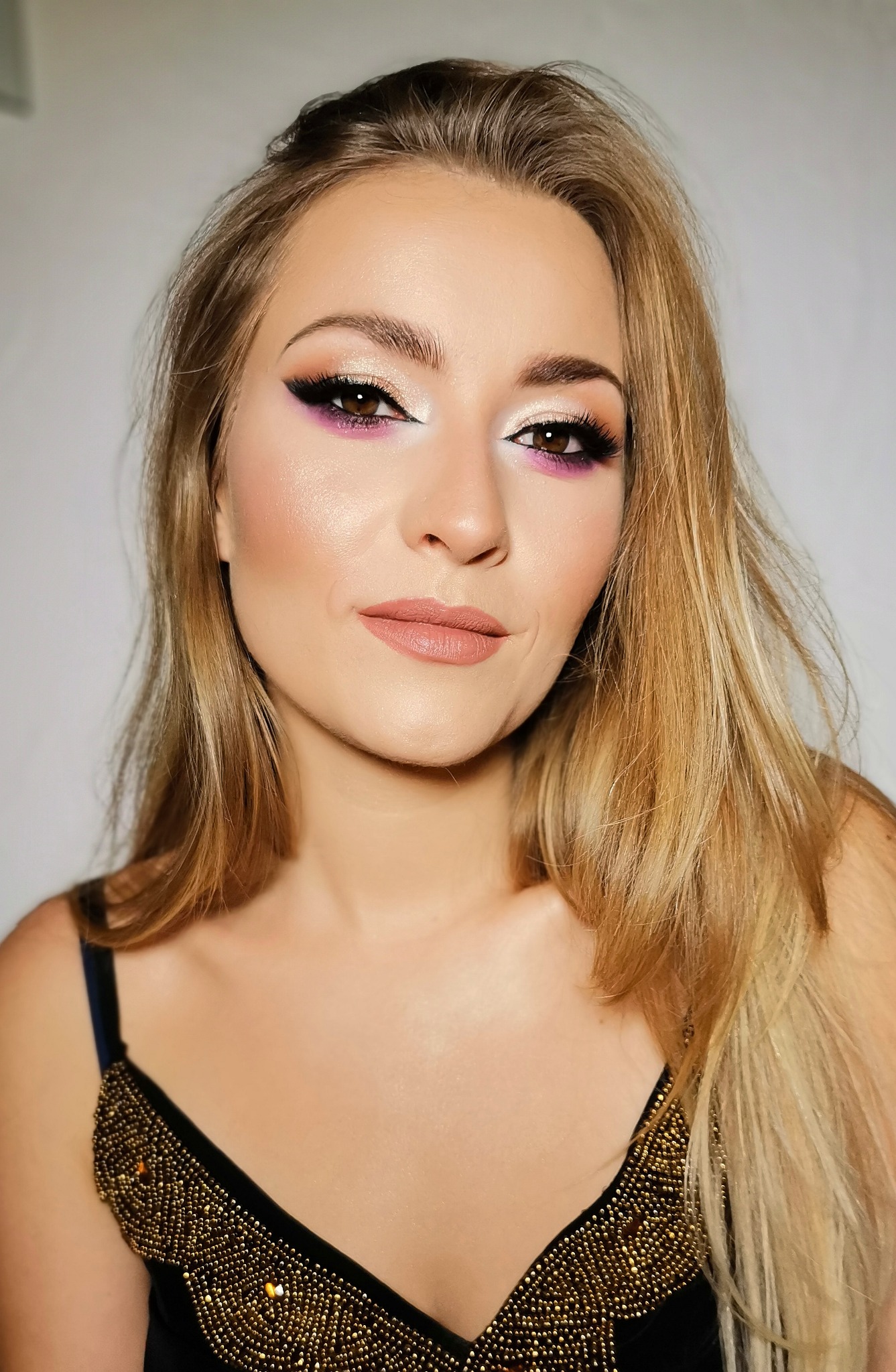 Portret kobiety z blond włosami i profesjonalnie wykonanym make-upem: fioletowe cienie na powiekach, podkreślone rzęsy i usta w odcieniu nude, ubrana w czarny top z ozdobnymi, złotymi koralikami.