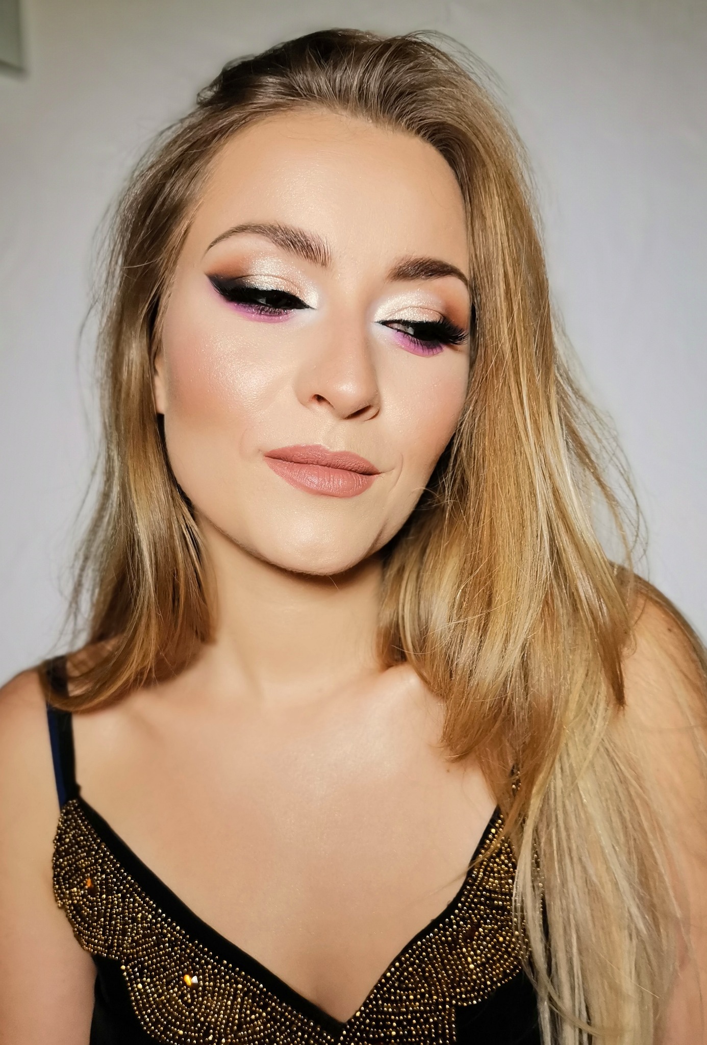 Portret kobiety z profesjonalnie wykonanym, wieczorowym wizażem, obejmującym złote cienie, czarny eyeliner i akcenty różu, w czarnej sukience z ozdobnymi, złotymi koralikami.