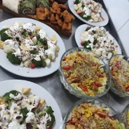 Różnorodne sałatki i przekąski ułożone na metalowym stole, obejmujące sałatki warzywne z brokułami i jajkiem, sałatki z kurczakiem i kukurydzą w szklanych misach oraz roladki i smażone kąski...