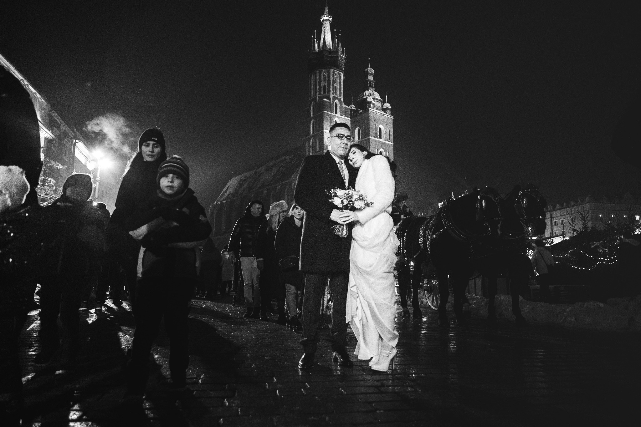 Czarno-białe zdjęcie pary młodej w Krakowie na tle Bazyliki Mariackiej, z dorożką konną i grupą ludzi w tle, nocna sceneria z mokrym brukiem.