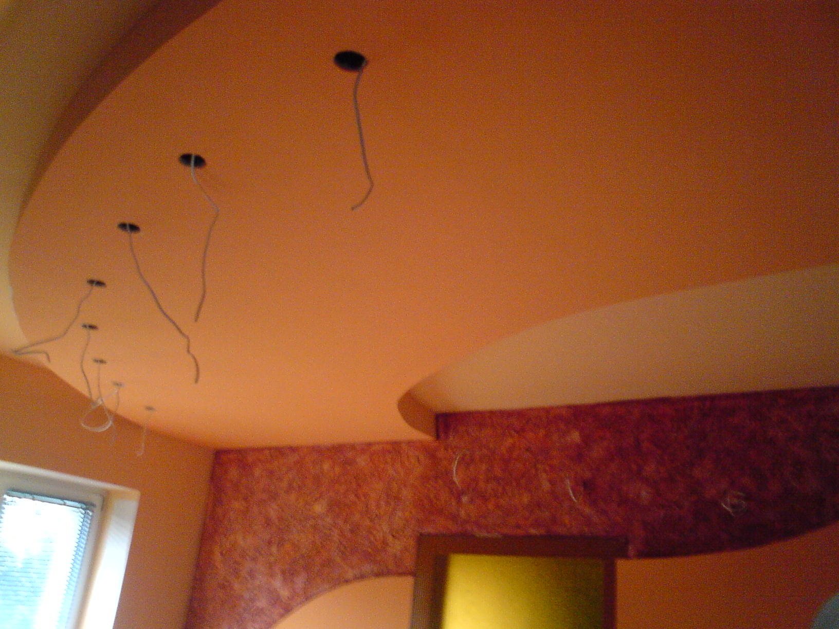 Pomarańczowe, zaokrąglone wykończenie sufitu podwieszanego z wystającymi przewodami elektrycznymi i widoczną ścianą z dekoracyjną fakturą.