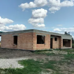 Surowy stan domu jednorodzinnego z czerwonej cegły, widoczne otwory okienne i drzwiowe, betonowy wieniec, w tle błękitne niebo z białymi chmurami, plac budowy.