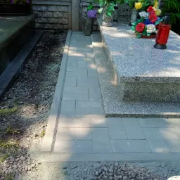 Ułożona szara kostka brukowa obok nagrobka z kwiatami i zniczem, widoczna świeżo położona podsypka z kamyczków.