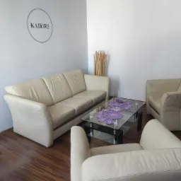 Jasny skórzany komplet wypoczynkowy: sofa i dwa fotele, na szklanym stoliku fioletowe serwety, na ścianie logo firmy KADORI.