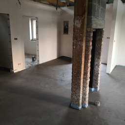 BRH Bracia Hudony usługi budowlane i wykończeniowe Maciej Hudon - Wnętrze w trakcie prac wykończeniowych: wylany betonowy podkład, odsłonięte ceglane filary z zabezpieczeniem, ściany przygotowane do malowania, widoczne instalacje elektryczne.