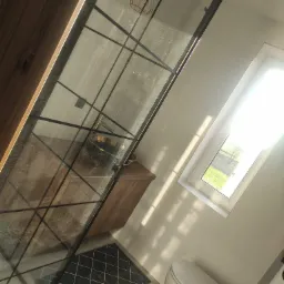 Nowoczesna łazienka z kabiną prysznicową typu walk-in z czarnymi szprosami, drewnianą szafką pod umywalką i widokiem na zieleń za oknem.