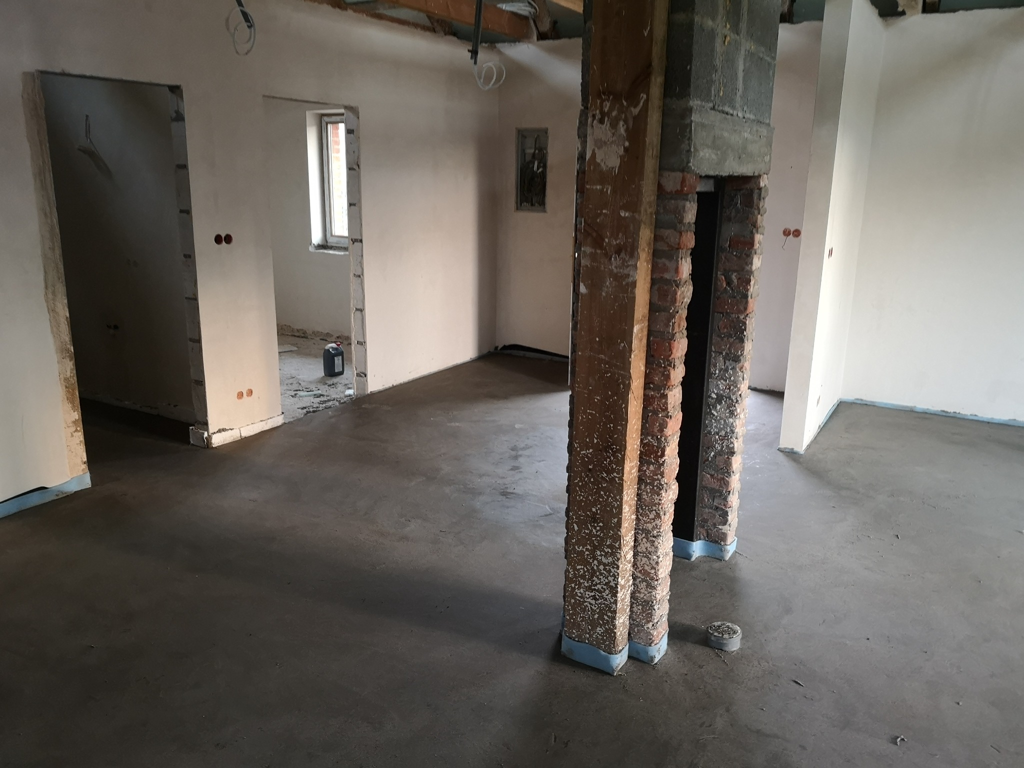 Wnętrze w trakcie prac wykończeniowych: wylany betonowy podkład, odsłonięte ceglane filary z zabezpieczeniem, ściany przygotowane do malowania, widoczne instalacje elektryczne.