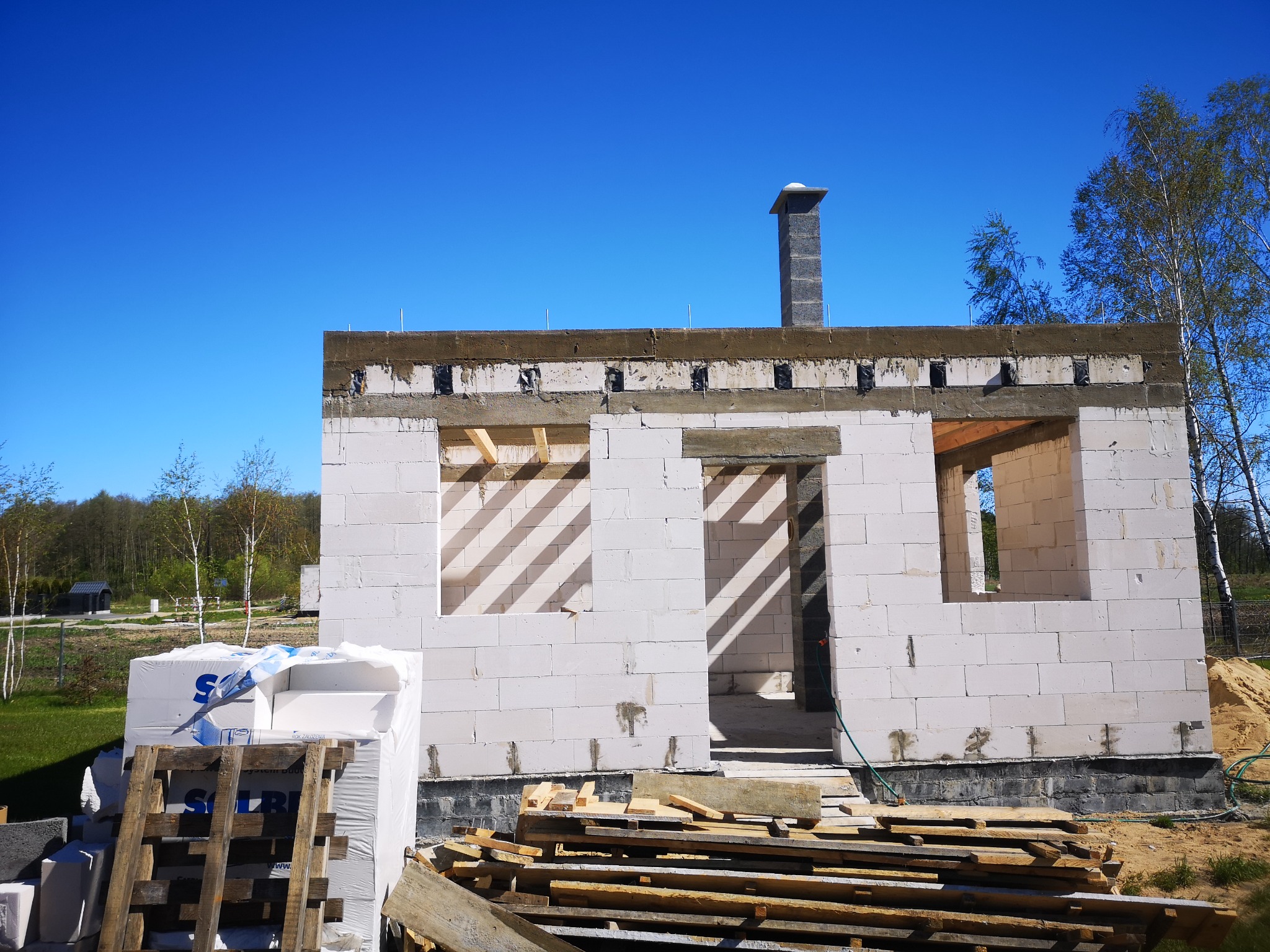 Surowy stan domu w trakcie budowy, ściany z białego betonu komórkowego, widoczne otwory okienne i komin, materiały budowlane składowane na paletach przed budynkiem, błękitne niebo w tle.