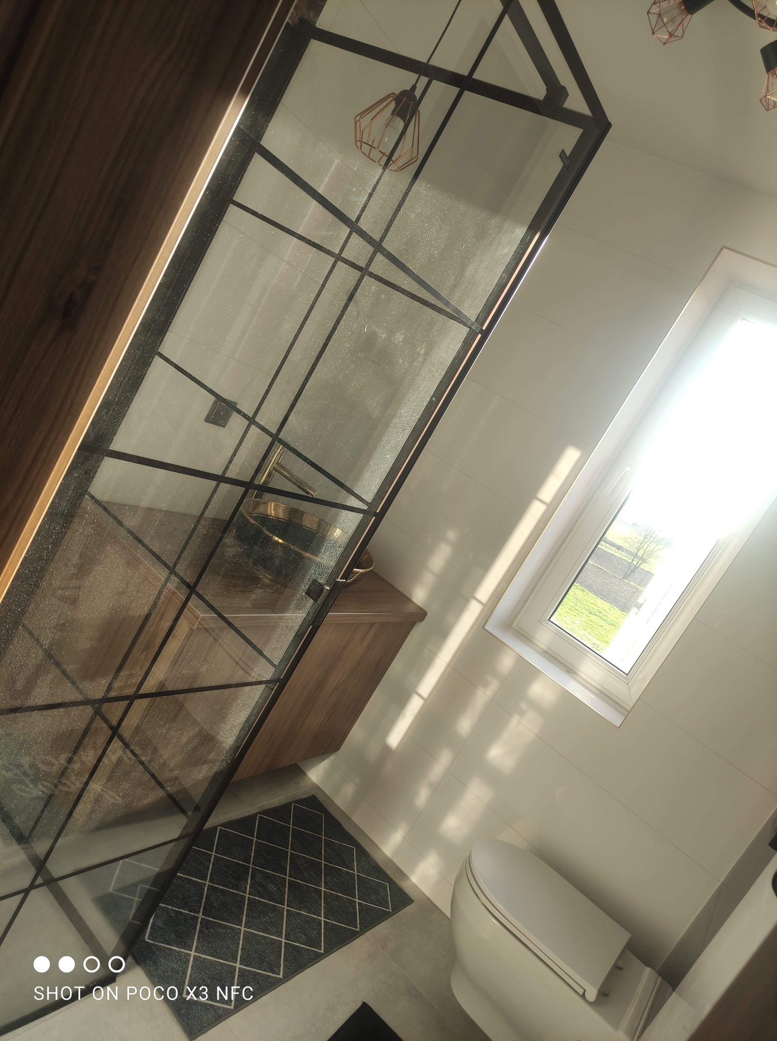 Nowoczesna łazienka z kabiną prysznicową typu walk-in z czarnymi szprosami, drewnianą szafką pod umywalką i widokiem na zieleń za oknem.