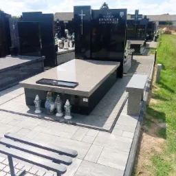 Nowoczesny, czarny pomnik z jasnoszarym blatem i ławką, ozdobiony lampionami, na cmentarzu w słoneczny dzień.