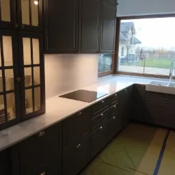 Szara kuchnia z białym blatem, widok na zewnątrz przez duże okno, zabezpieczona podłoga folią malarską.